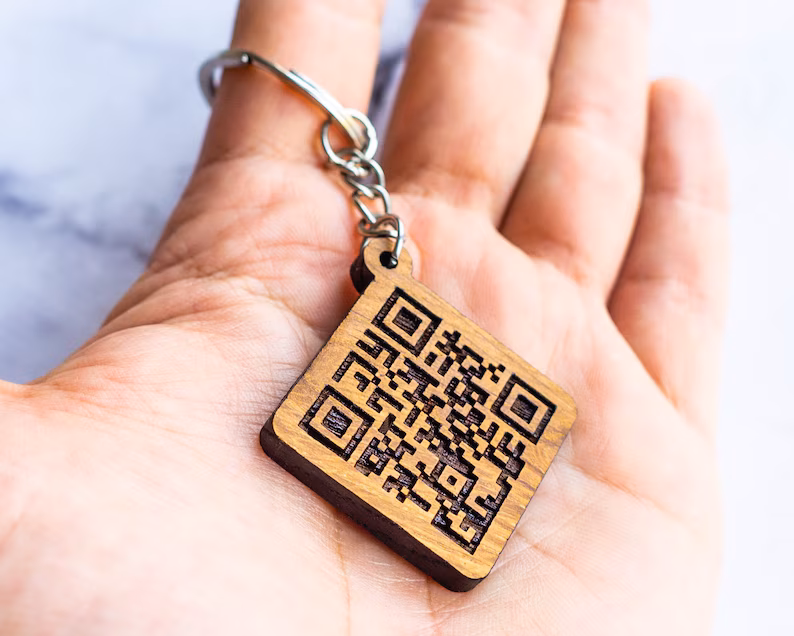 Eine Handfläche die einen Schlüsselanhänger aus Holz trägt auf dem ein QR Code gelasert ist.